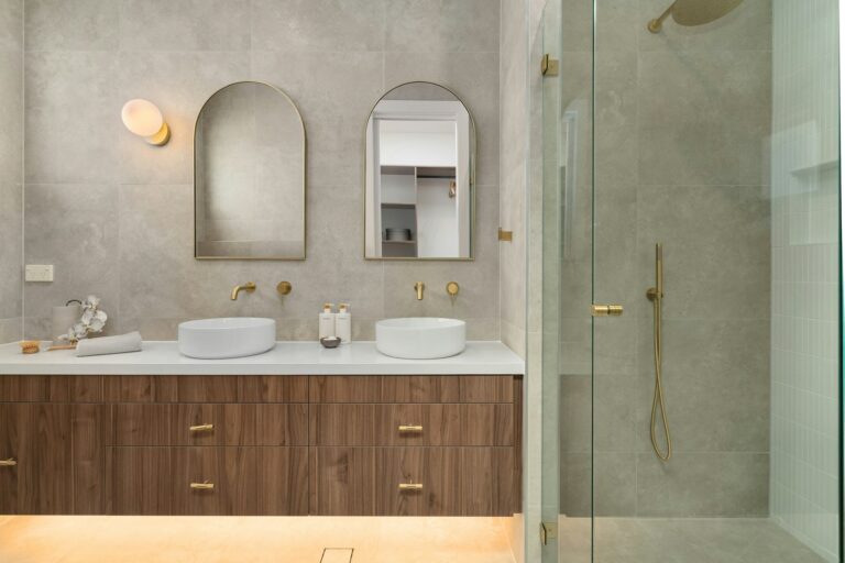 Warm stone bathroom with a wall-hung walnut vanity, brushed brass fixtures, arched mirrors and a walk-in glass shower