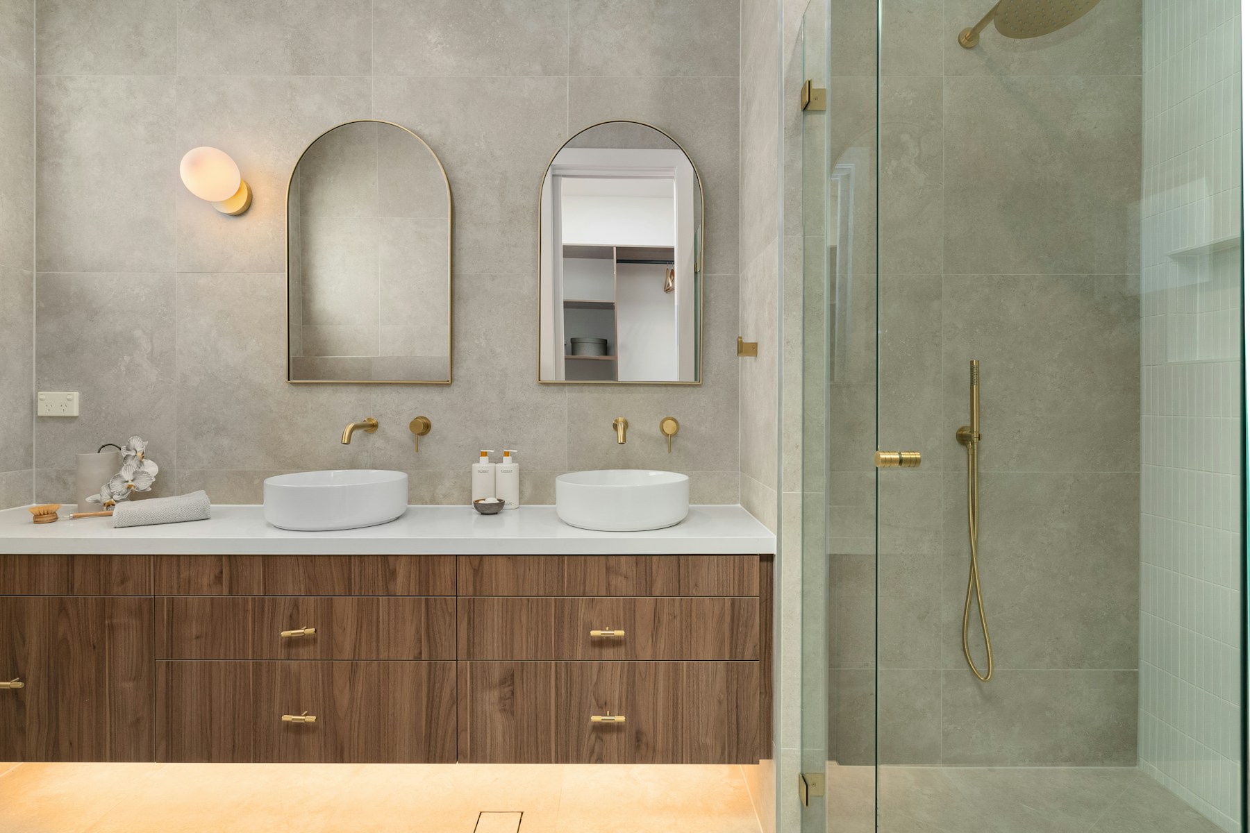 Warm stone bathroom with a wall-hung walnut vanity, brushed brass fixtures, arched mirrors and a walk-in glass shower